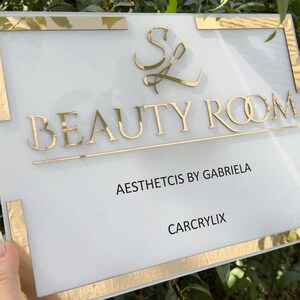 May include: A rectangular sign with a white background and gold trim. The sign reads "BEAUTY ROOM" in gold lettering, with additional text below. The sign also features a gold logo and the text "AESTHETICS BY GABRIELA" and "CARCRYLIX".