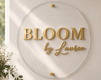 Puede incluir: Un letrero circular de acrílico transparente con letras en relieve de color dorado que dicen "BLOOM by Lauren". El letrero está montado con dos pequeñas piezas de hardware circulares de color dorado en el centro superior e inferior.