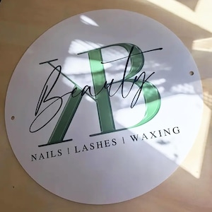May include: A circular white sign featuring the word "Beauty" in elegant cursive script above a green "AB" design. The text "NAILS | LASHES | WAXING" is printed below. Two small holes are visible on the sign's edge.