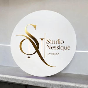 May include: A circular white sign featuring gold lettering and a gold logo. The sign displays "Studio Nessique BY RESSA". The logo is an abstract design incorporating the letters "S" and "N".