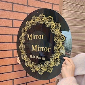 May include: A circular black sign featuring gold decorative detailing and the words "Mirror Mirror Hair Design" in gold. The sign is mounted on a brick wall.
