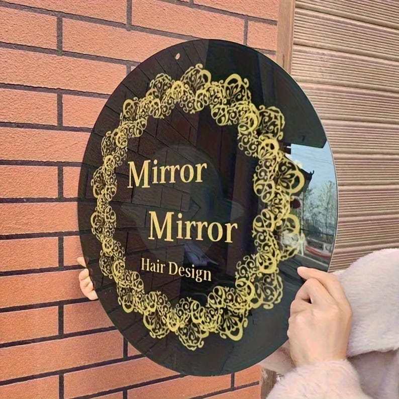 May include: A circular black sign featuring gold decorative detailing and the words "Mirror Mirror Hair Design" in gold. The sign is mounted on a brick wall.