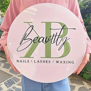 May include: A circular, pink sign featuring the word "Beauty" in cursive script, with the letters "KB" in a green, stylized font. The sign also displays the words "NAILS | LASHES | WAXING" in a black, sans-serif font.