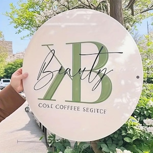 Puede incluir: Un letrero redondo blanco con la palabra "Beauty" en escritura cursiva y las letras "KB" en verde. El letrero también incluye el texto "COSE COFFEE SEGITCE". El diámetro del letrero es de 60 cm.