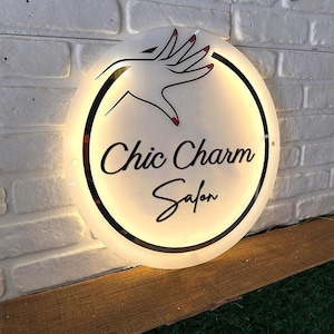 Può includere: Un'insegna rotonda e illuminata con la scritta "Chic Charm Salon" in elegante corsivo. L'insegna presenta un contorno nero e una grafica di una mano con smalto rosso. È montata su un muro di mattoni bianchi.