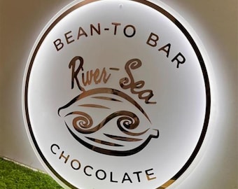 May include: A round, illuminated sign with the words "BEAN-TO BAR CHOCOLATE" in a circular arrangement. The sign features the text "River-Sea" in a decorative font, along with a stylized cocoa bean and wave design.