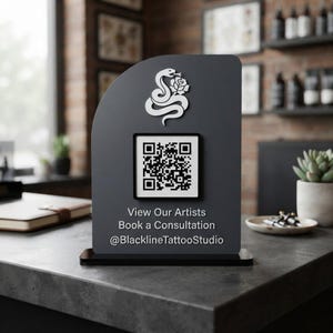 May include: A dark gray, arched sign featuring a white snake and rose graphic, a central QR code, and the text: View Our Artists, Book a Consultation, @BlacklineTattooStudio. The sign is mounted on a black rectangular base for display.