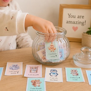 Peut inclure: Un bocal en verre transparent rempli de cartes colorées avec des illustrations d'animaux et des affirmations positives. Les cartes sont disposées sur une surface en bois. Une main d'enfant atteint le bocal. Un panneau encadré indique "You are amazing!"