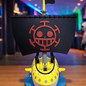 Może przedstawiać: Żółty model łodzi podwodnej z czarnym żaglem z czerwonym emblematem. Model ma biały przód i stoi na niebieskiej podstawie. Czarna flaga znajduje się na maszcie.