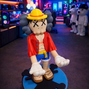 Może przedstawiać: Kolorowa figurka postaci z kreskówki w żółtym kapeluszu, czerwonej kurtce i niebieskich szortach. Postać ma białe rękawiczki i sandały, stojąc na niebieskiej podstawie z motywem czaszki. Tło przedstawia pokój z komputerami i kolorowymi światłami.