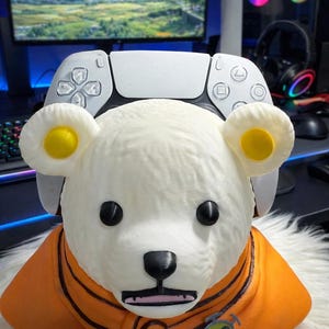 Może przedstawiać: Biała figurka w kształcie niedźwiedzia z czarnymi oczami i nosem, ubrana w pomarańczową kurtkę z zielono-żółtym emblematem. Biały kontroler do gier znajduje się na głowie niedźwiedzia. Tło obejmuje monitor komputera i sprzęt do gier.