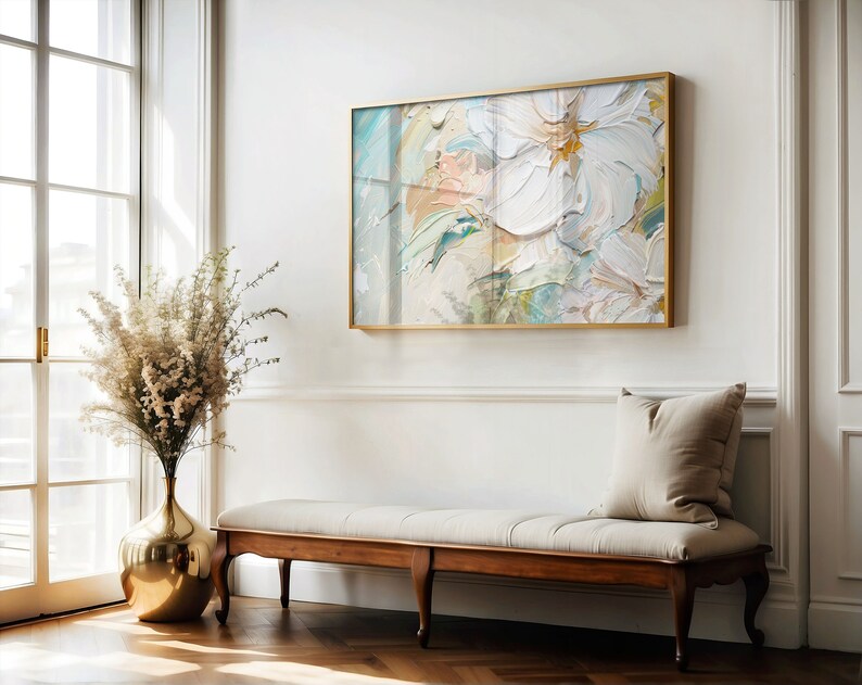 May include: A white, gold, and blue abstract painting in a gold frame hangs on a white wall above a wooden bench with a white cushion and a beige pillow. A gold vase with white flowers sits on the floor next to the bench.