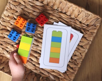 May include: A child's hand holds a stack of colorful Lego blocks. The blocks are orange, yellow, green, and blue. There are also several other Lego blocks scattered on a woven basket. There are three cards with images of Lego blocks on them.