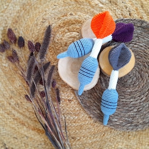 May include: Three crocheted teethers in the shape of a dart. The teethers are blue, orange, and gray. The teethers are on a woven background.