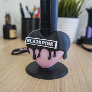 May include: A black heart-shaped phone stand with a black label that says "BLACKPINK" in white letters. The stand has a pink heart-shaped base.
