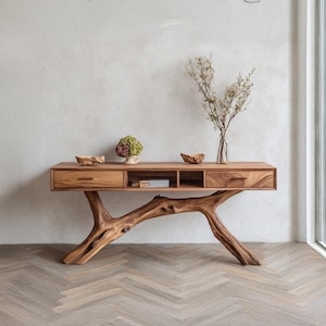 Peut inclure: Une console en bois avec une base en branche d'arbre naturelle. La table comporte deux tiroirs et des étagères ouvertes. Des objets décoratifs, dont un petit vase de fleurs et des bols en bois, sont exposés sur la surface de la table. La table est en bois brun clair.