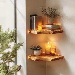 Puede incluir: Dos estanterías de esquina de madera con libros, plantas y velas. La estantería superior contiene libros, un jarrón con vegetación y una vela pequeña. La estantería inferior tiene una planta pequeña, una vela grande y un portavelas decorativo. Luces de cadena envueltas alrededor de los bordes.