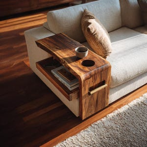 Puede incluir: Una mesa auxiliar de madera con un diseño de borde natural. La mesa tiene un portavasos incorporado y un estante inferior. Una pequeña taza de cerámica está en el portavasos. La mesa está junto a un sofá beige con un cojín marrón.