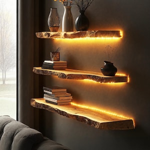 May include: Three wooden shelves with a natural wood finish and a rough-hewn edge. The shelves are lit with warm white LED lights. The top shelf has a vase with dried flowers, a black vase, and a glass lantern. The middle shelf has a stack of books and a small black vase. The bottom shelf has a stack of books.
