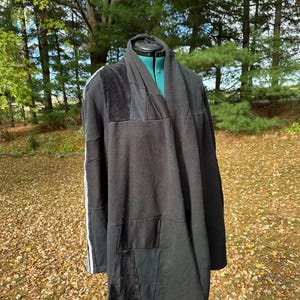 May include: A black patchwork cardigan with a draped collar. The cardigan features a mix of textures, including fleece and faux fur, and has white stripes down the sleeve. The garment is displayed on a mannequin outdoors.