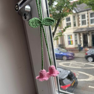 May include: A crocheted flower charm with pink flowers and green leaves hanging from a window handle.
