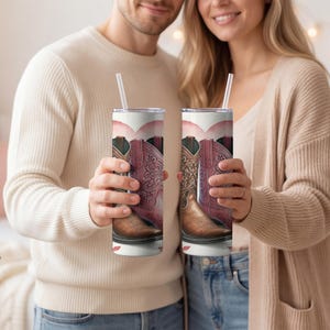 Op de afbeelding: Twee cilindrische roestvrijstalen bekers met een grafische print van bruine en roze cowboylaarzen tegen een zachte hartvormige achtergrond. Elke beker is voorzien van een doorzichtig deksel en een wit herbruikbaar rietje.