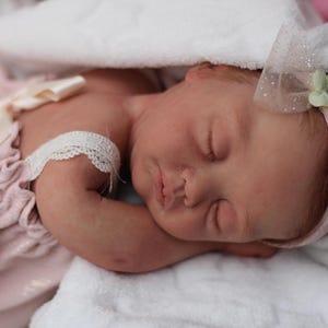 Puede incluir: Una mu&ntilde;eca beb&eacute; durmiendo con un vestido rosa con ribete de encaje y una diadema rosa con una flor blanca.
