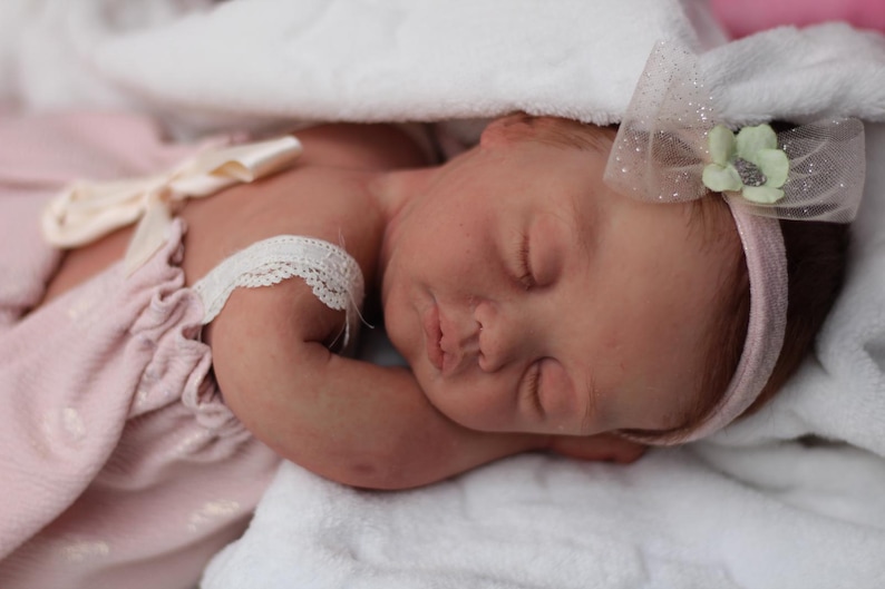 Puede incluir: Una mu&ntilde;eca beb&eacute; durmiendo con un vestido rosa con ribete de encaje y una diadema rosa con una flor blanca.