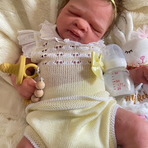 May include: A lifelike baby doll wearing a yellow knitted romper with white trim and a matching headband with a bow. The doll has a pacifier and a bottle with measurements. A white stuffed bunny is in the background.