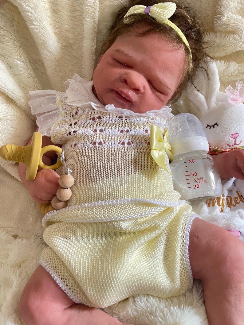 May include: A lifelike baby doll wearing a yellow knitted romper with white trim and a matching headband with a bow. The doll has a pacifier and a bottle with measurements. A white stuffed bunny is in the background.