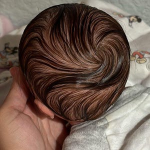 Puede incluir: Primer plano de la cabeza de una mu&ntilde;eca beb&eacute; con cabello enraizado marr&oacute;n peinado en espiral.
