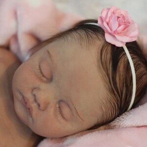 Puede incluir: Una mu&ntilde;eca beb&eacute; durmiendo con cabello casta&ntilde;o que lleva una diadema de flores rosa. La mu&ntilde;eca est&aacute; acostada sobre una manta rosa con lunares blancos.