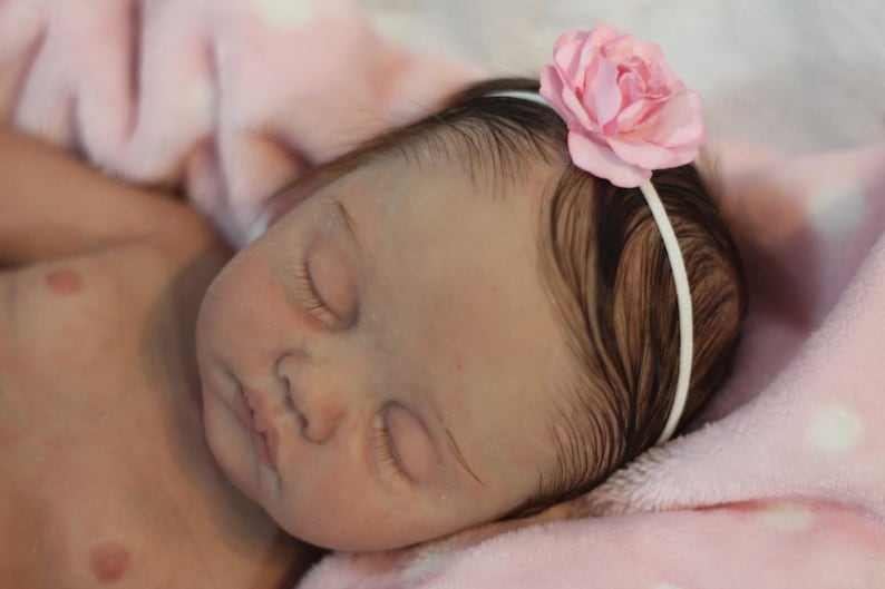 Puede incluir: Una mu&ntilde;eca beb&eacute; durmiendo con cabello casta&ntilde;o que lleva una diadema de flores rosa. La mu&ntilde;eca est&aacute; acostada sobre una manta rosa con lunares blancos.