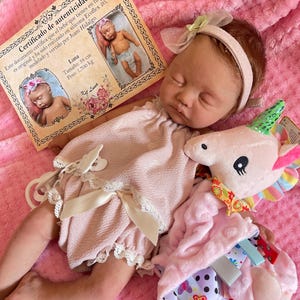 Puede incluir: Una mu&ntilde;eca de beb&eacute; rosa y blanca con cabello casta&ntilde;o y ojos cerrados est&aacute; acostada sobre una manta rosa. La mu&ntilde;eca lleva un vestido rosa con ribete de encaje blanco y una diadema blanca con un lazo rosa. La mu&ntilde;eca est&aacute; sosteniendo un animal de peluche unicornio rosa y verde.