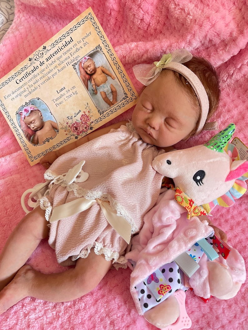 Puede incluir: Una mu&ntilde;eca de beb&eacute; rosa y blanca con cabello casta&ntilde;o y ojos cerrados est&aacute; acostada sobre una manta rosa. La mu&ntilde;eca lleva un vestido rosa con ribete de encaje blanco y una diadema blanca con un lazo rosa. La mu&ntilde;eca est&aacute; sosteniendo un animal de peluche unicornio rosa y verde.
