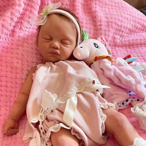 Puede incluir: Una mu&ntilde;eca de beb&eacute; rosa y blanca con un juguete de peluche de unicornio. La mu&ntilde;eca lleva un vestido rosa con ribete de encaje blanco y una diadema blanca con un lazo verde. La mu&ntilde;eca tiene los ojos cerrados y una apariencia realista.