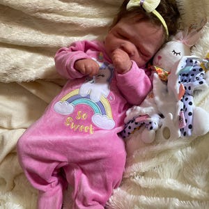 May include: A sleeping baby doll wearing a pink onesie with a unicorn and rainbow design, the text "So Sweet", and a yellow bow headband. A white stuffed bunny with a pink bow and polka dot accents rests beside the doll.