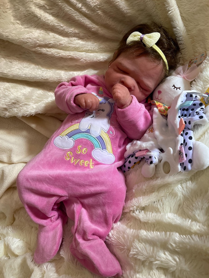 May include: A sleeping baby doll wearing a pink onesie with a unicorn and rainbow design, the text "So Sweet", and a yellow bow headband. A white stuffed bunny with a pink bow and polka dot accents rests beside the doll.