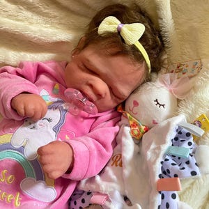 May include: A sleeping baby doll wearing a pink onesie with a unicorn graphic and the text "So Sweet". The doll has a yellow bow in its hair and a pacifier in its mouth. A white plush bunny toy is next to the doll.