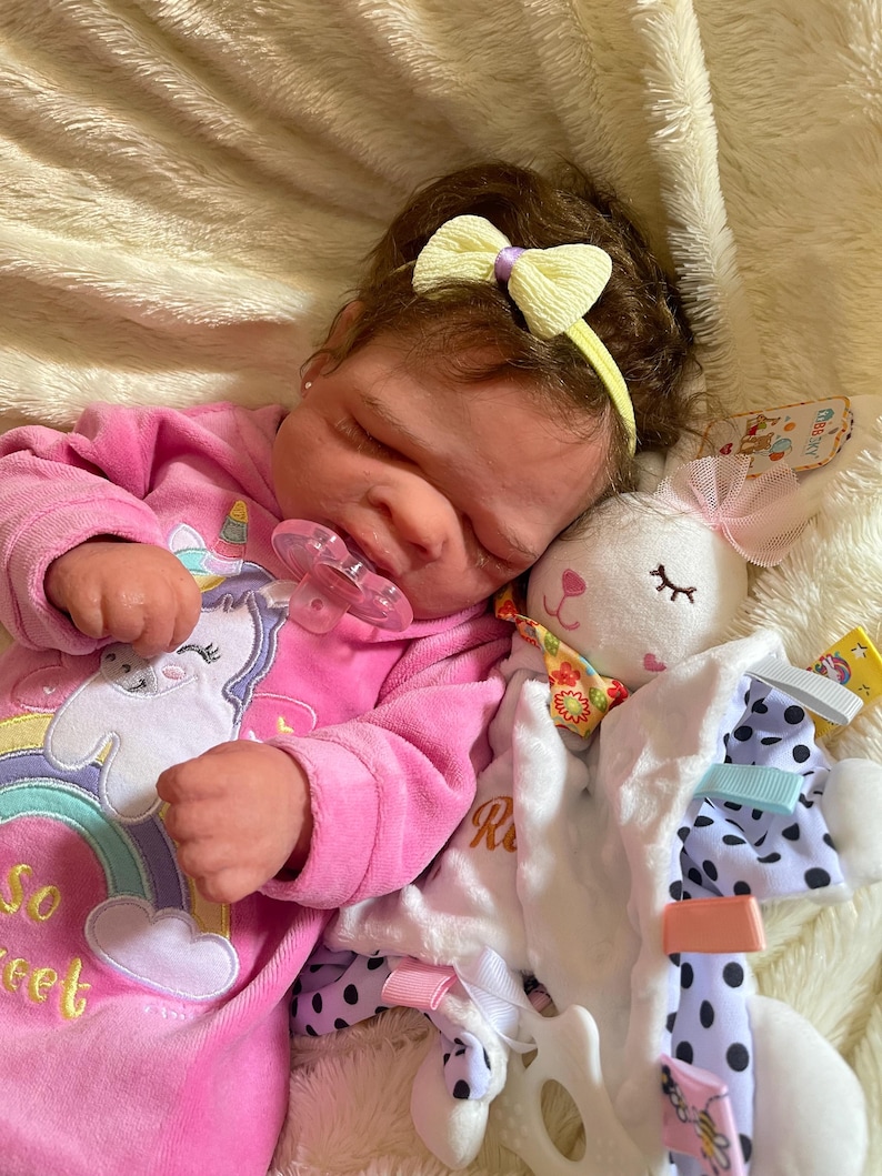 May include: A sleeping baby doll wearing a pink onesie with a unicorn graphic and the text "So Sweet". The doll has a yellow bow in its hair and a pacifier in its mouth. A white plush bunny toy is next to the doll.