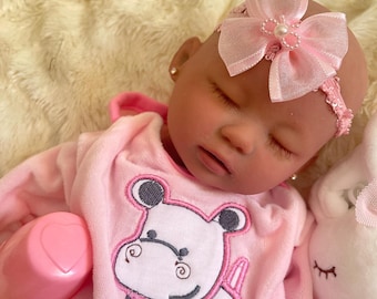 May include: A pink and white baby doll with closed eyes, wearing a pink and white outfit with a hippopotamus design. The doll is wearing a pink headband with a bow and a pearl.