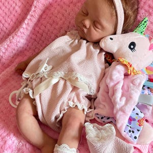 Puede incluir: Una mu&ntilde;eca de beb&eacute; rosa y blanca con cabello casta&ntilde;o lleva un vestido rosa y calcetines blancos. La mu&ntilde;eca sostiene un juguete de peluche rosa y blanco en forma de unicornio.