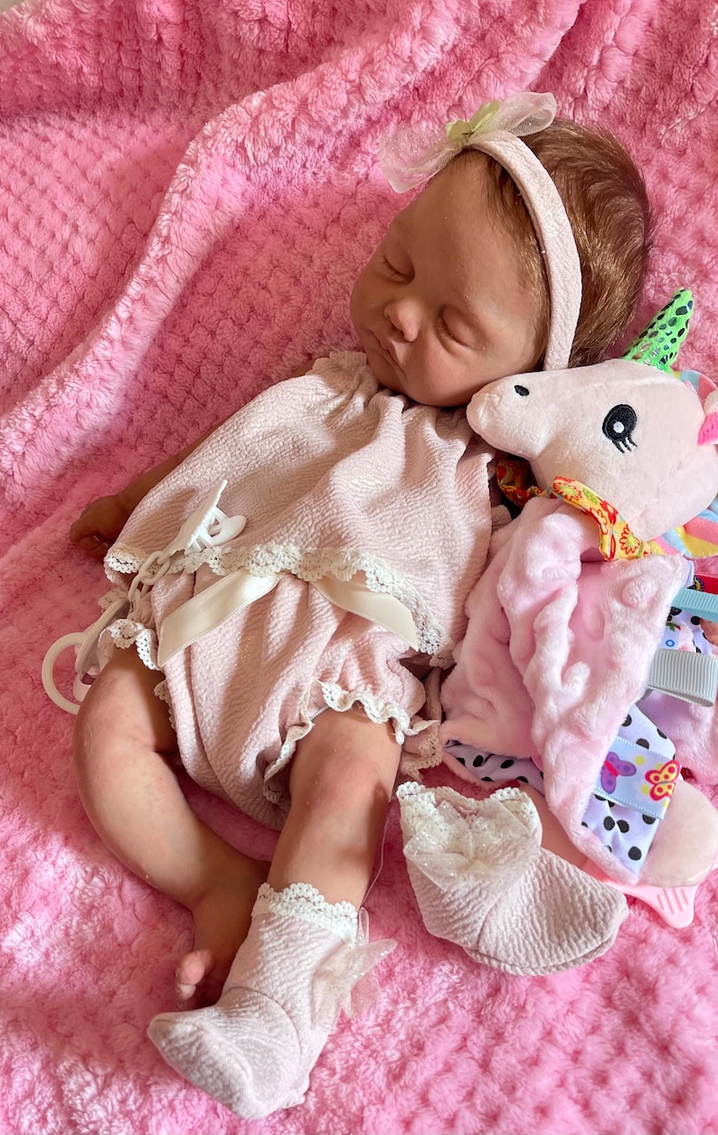 Puede incluir: Una mu&ntilde;eca de beb&eacute; rosa y blanca con cabello casta&ntilde;o lleva un vestido rosa y calcetines blancos. La mu&ntilde;eca sostiene un juguete de peluche rosa y blanco en forma de unicornio.