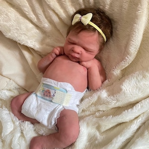 May include: A lifelike baby doll with brown hair and a yellow bow headband. The doll is wearing a diaper and is lying on a soft, white blanket. The doll's skin has a realistic tone.