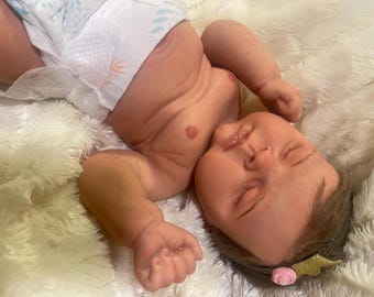 Puede incluir: Una muñeca bebé realista con los ojos cerrados, que lleva un pañal. La muñeca tiene una cara detallada con rasgos pintados y una diadema con una flor rosa y alas doradas. La muñeca está tumbada sobre una manta blanca y esponjosa.