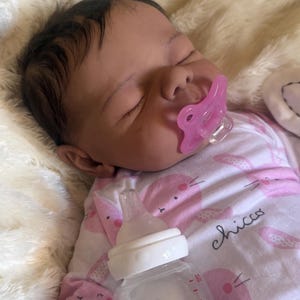 Puede incluir: Una muñeca realista con cabello oscuro y ojos cerrados, que lleva un body blanco con un patrón de conejitos rosas y la palabra chicco impresa. La muñeca tiene un chupete rosa en la boca y está colocada junto a un biberón de plástico transparente.