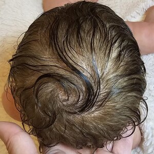 May include: Close-up of a baby doll's head with brown, wet-looking hair. The hair is styled in loose curls and waves, with a natural color gradient. The doll's head is resting on a soft, white surface.