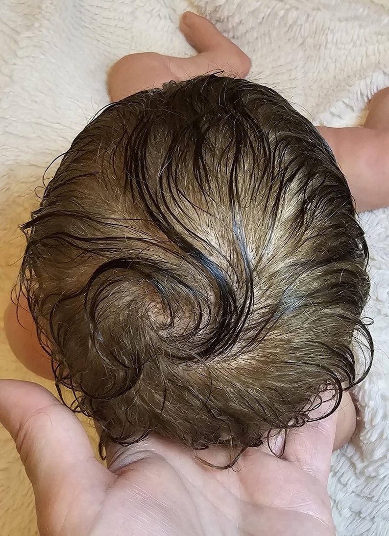 May include: Close-up of a baby doll's head with brown, wet-looking hair. The hair is styled in loose curls and waves, with a natural color gradient. The doll's head is resting on a soft, white surface.