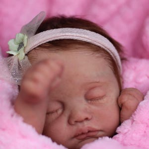 Puede incluir: Un mu&ntilde;eco beb&eacute; durmiendo con cabello casta&ntilde;o que lleva una diadema blanca con un adorno de flor verde. El mu&ntilde;eco est&aacute; acostado sobre una manta rosa peluda.