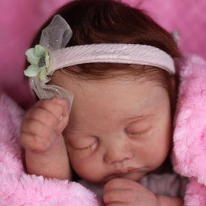 Puede incluir: Un mu&ntilde;eco beb&eacute; durmiendo con una diadema rosa y una flor verde. El mu&ntilde;eco lleva un vestido blanco y est&aacute; acostado en una manta rosa.